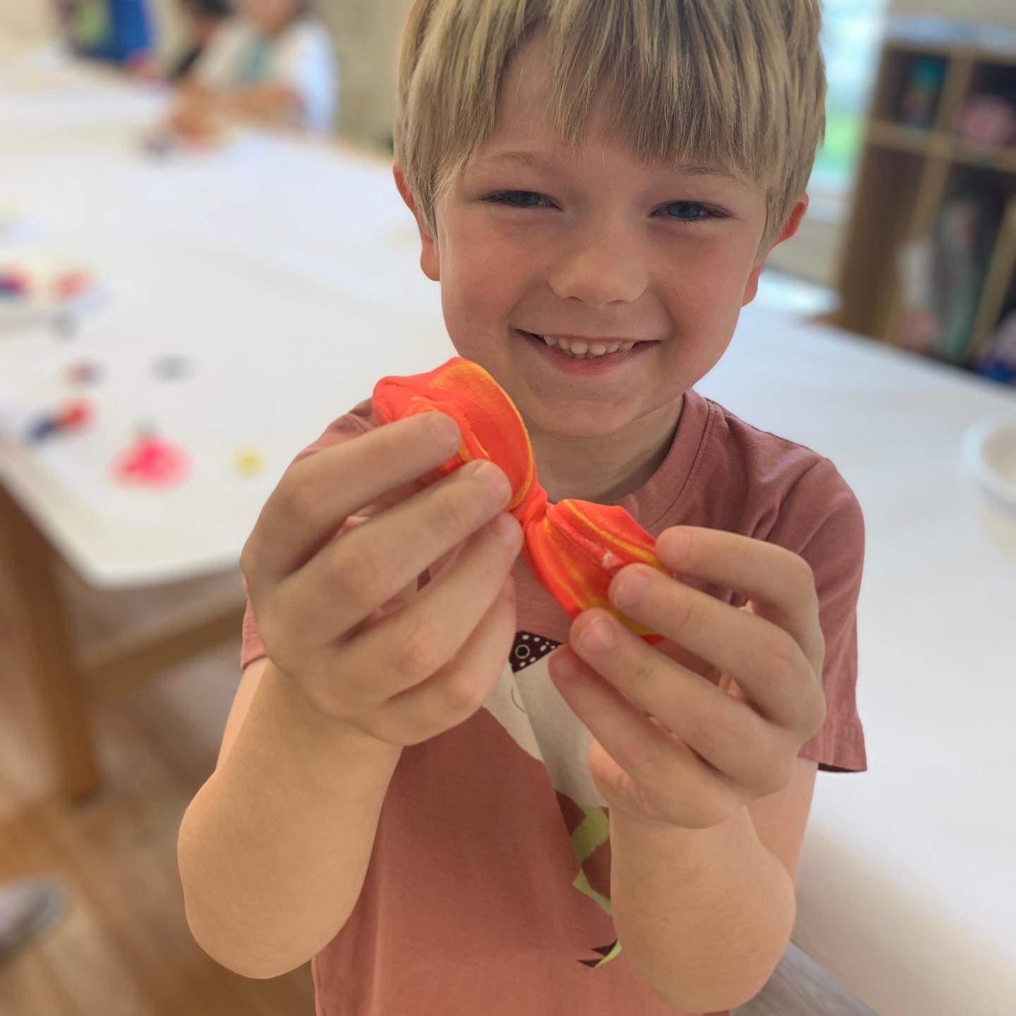 A young boy smiles, holding up an orange paper bow in a classroom setting.