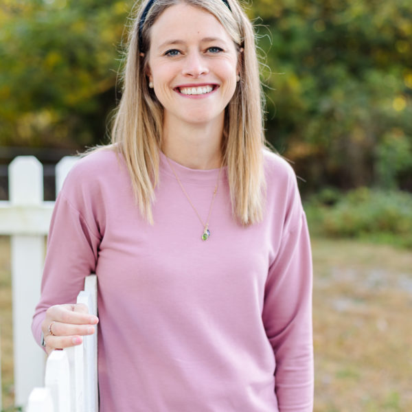 Blonde woman in pink top smiles, leaning on a white picket fence outdoors.