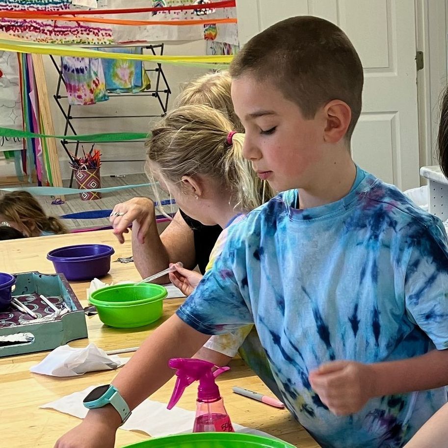 Two children in a classroom proudly display their colorful artwork. A boy smiles, holding a drawing of a volcano and an alien ship. A girl smiles with her mouth open and holds another art piece.