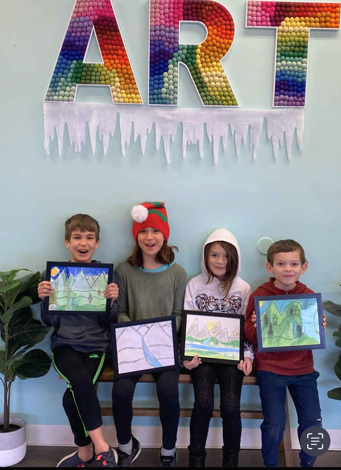 Four children seated, holding their artwork, smiling in front of a colorful “ART” sign with icicles, inside an art studio.