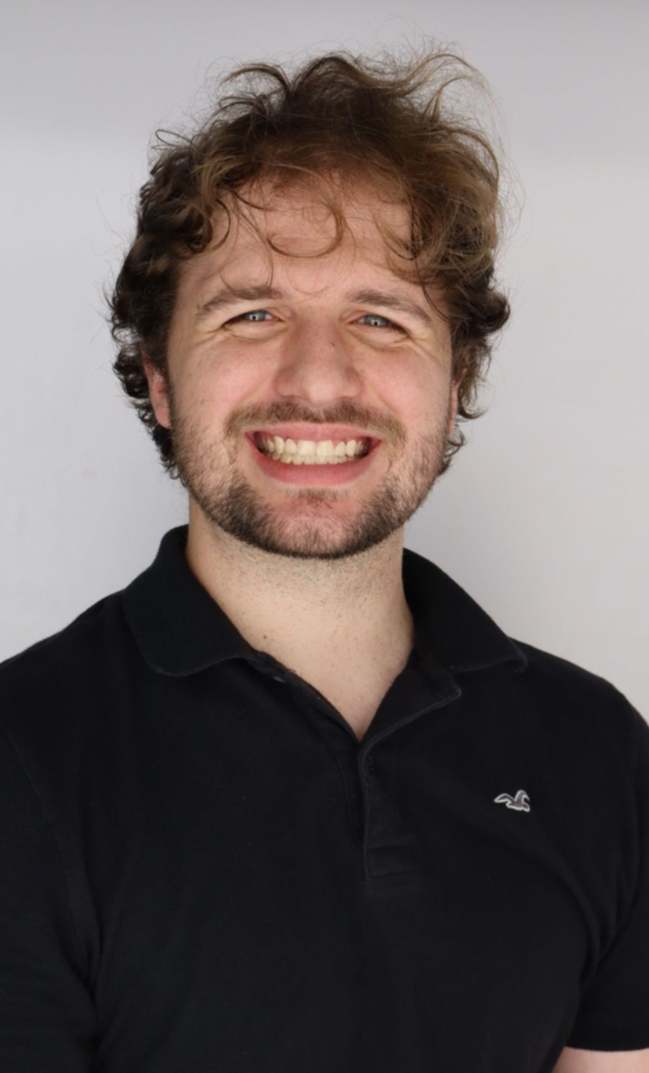 Man with curly brown hair and a beard smiles broadly, wearing a black polo shirt against a white background.