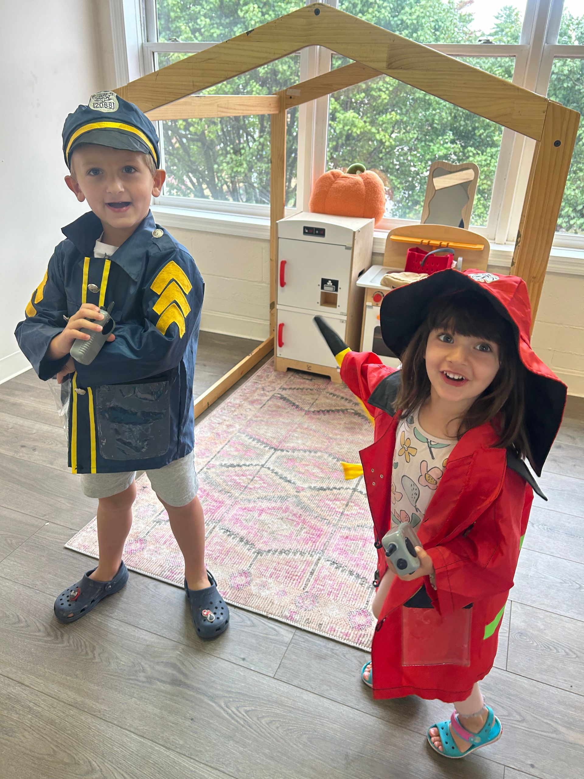 Two young children dressed as a police officer and firefighter smiling in front of a playhouse.