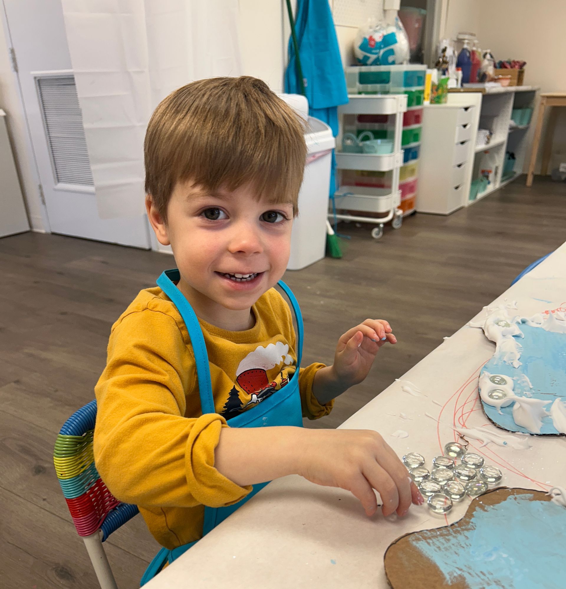 Young child in a yellow shirt and blue apron smiles while crafting, with craft supplies visible in the background.