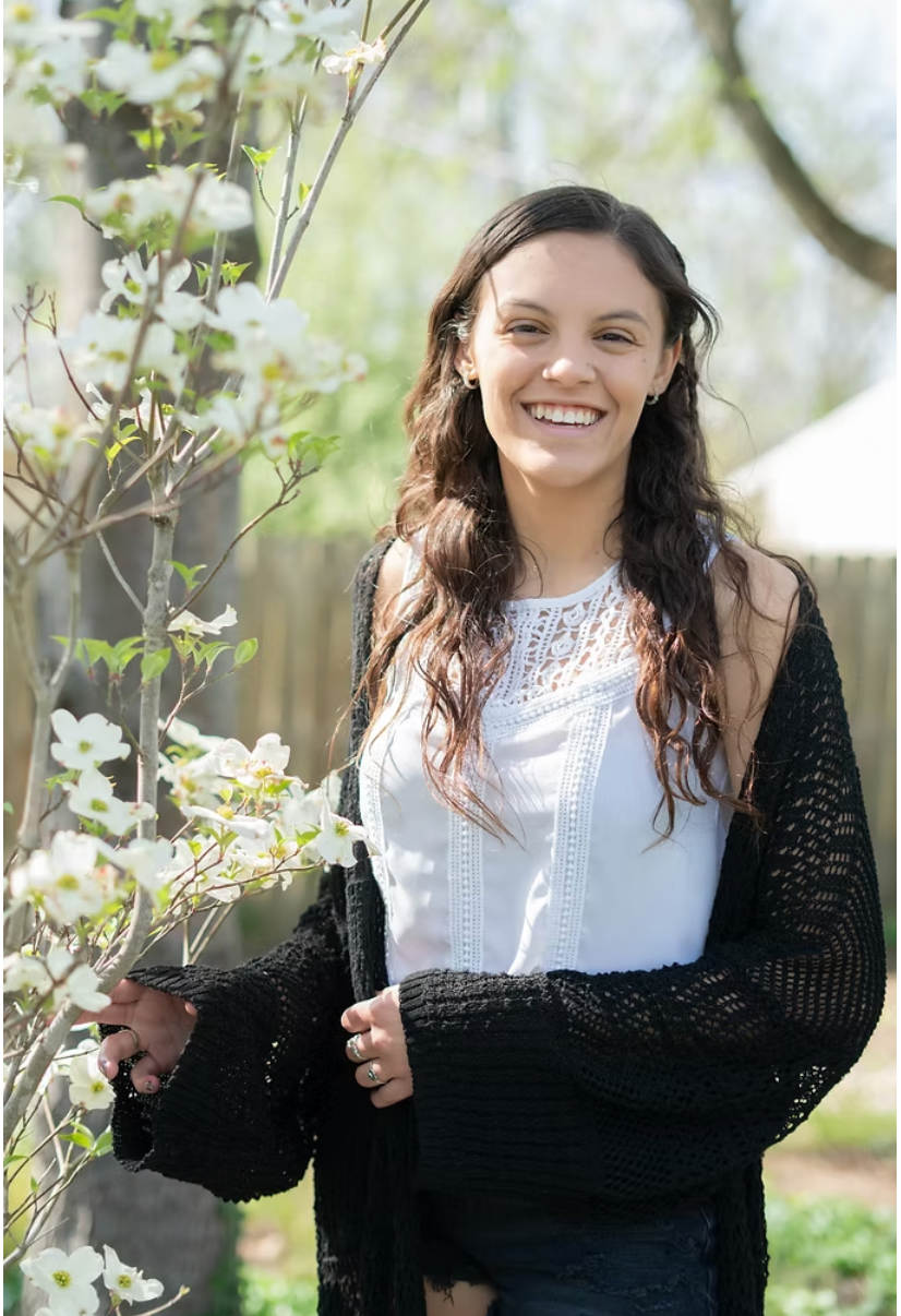 A young woman with long brown hair smiles, wearing a white top and black cardigan, standing by a flowering tree.