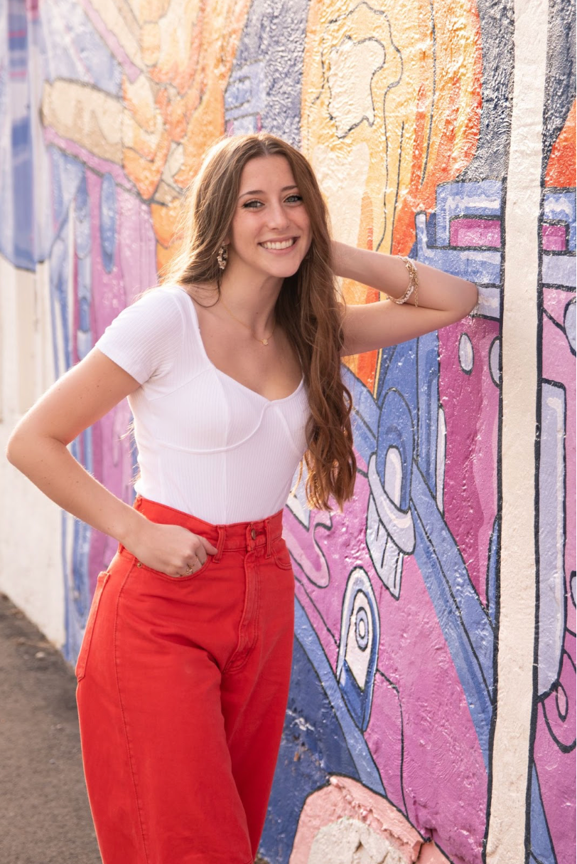 Young woman smiles while leaning against a colorful mural, wearing a white top and red pants.
