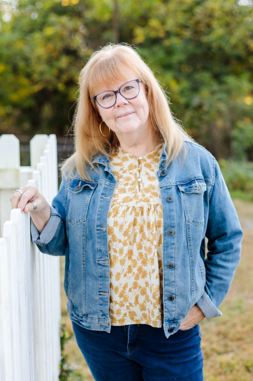 Woman with blond hair and glasses, wearing a denim jacket, and leaning against a white picket fence, smiling outdoors.