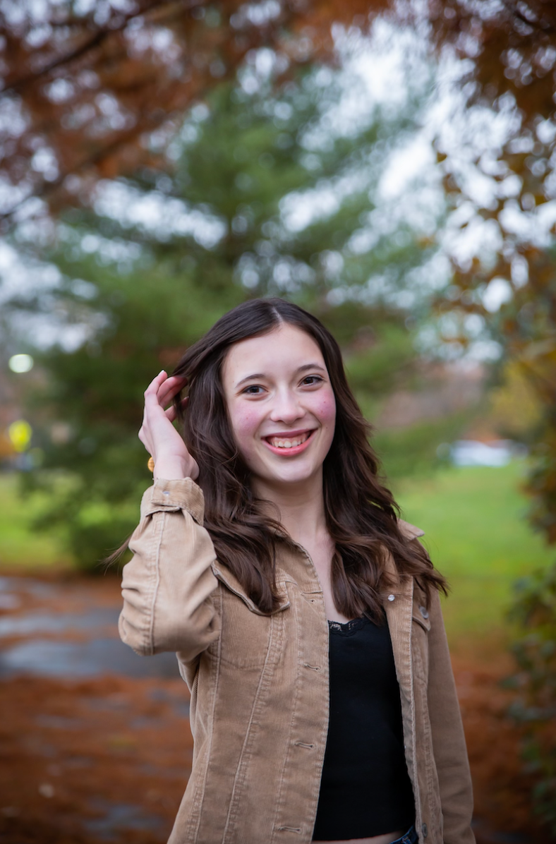 A young woman with braces smiles while holding her hair, wearing a beige jacket and black shirt, outdoors in a park.