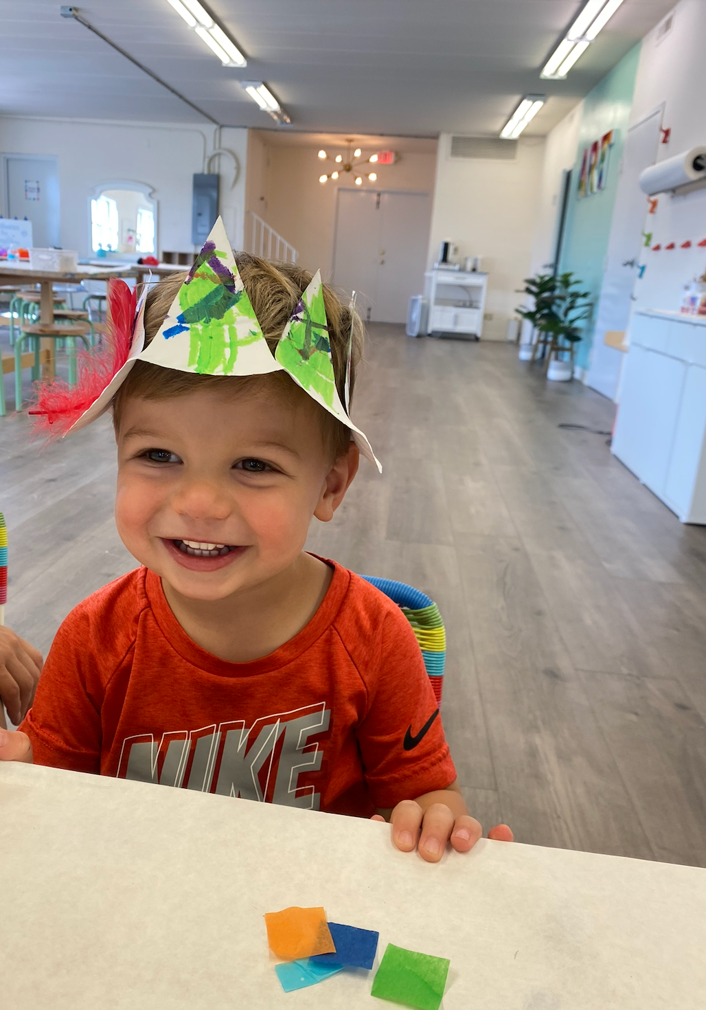 Smiling child wearing a decorated paper crown, seated at a table in a brightly lit classroom.