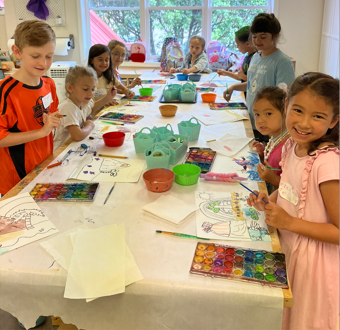 Children are painting at a table in a brightly lit classroom. Watercolor palettes, paper, and art supplies are spread out.