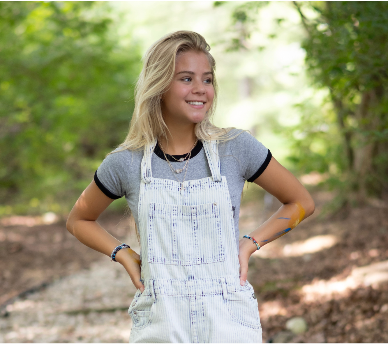 Blonde teen girl wearing overalls smiles in a forest setting. She has her hands on her hips.