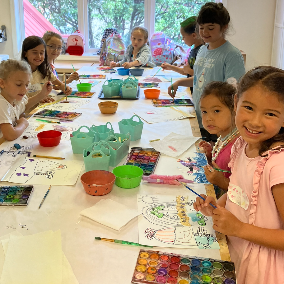 Children painting at a long table in a well-lit room. Colorful art supplies and smiling faces are visible.