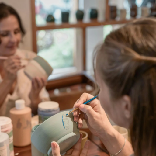 Two women paint pottery at a table indoors. One focuses on the pottery's detail, while the other paints a pot in the background.