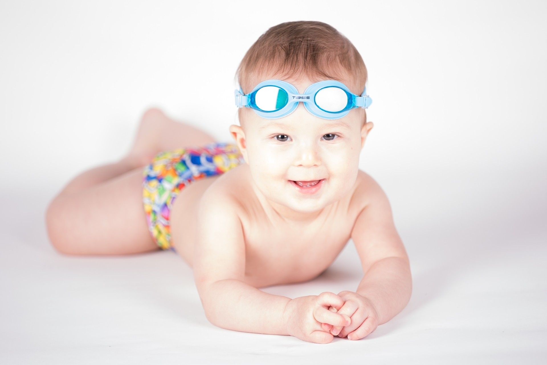 A baby wearing swimming goggles is laying on his stomach on a white surface.