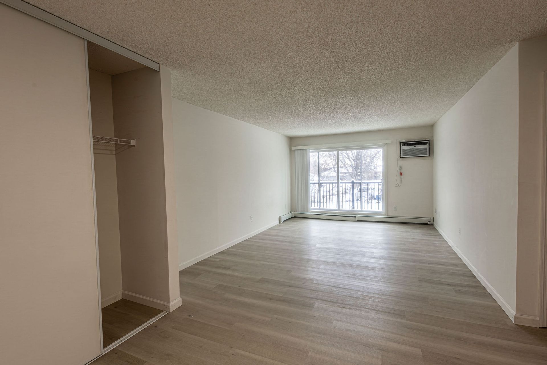Empty apartment living room with wood floors, white walls, and large windows at the far end
