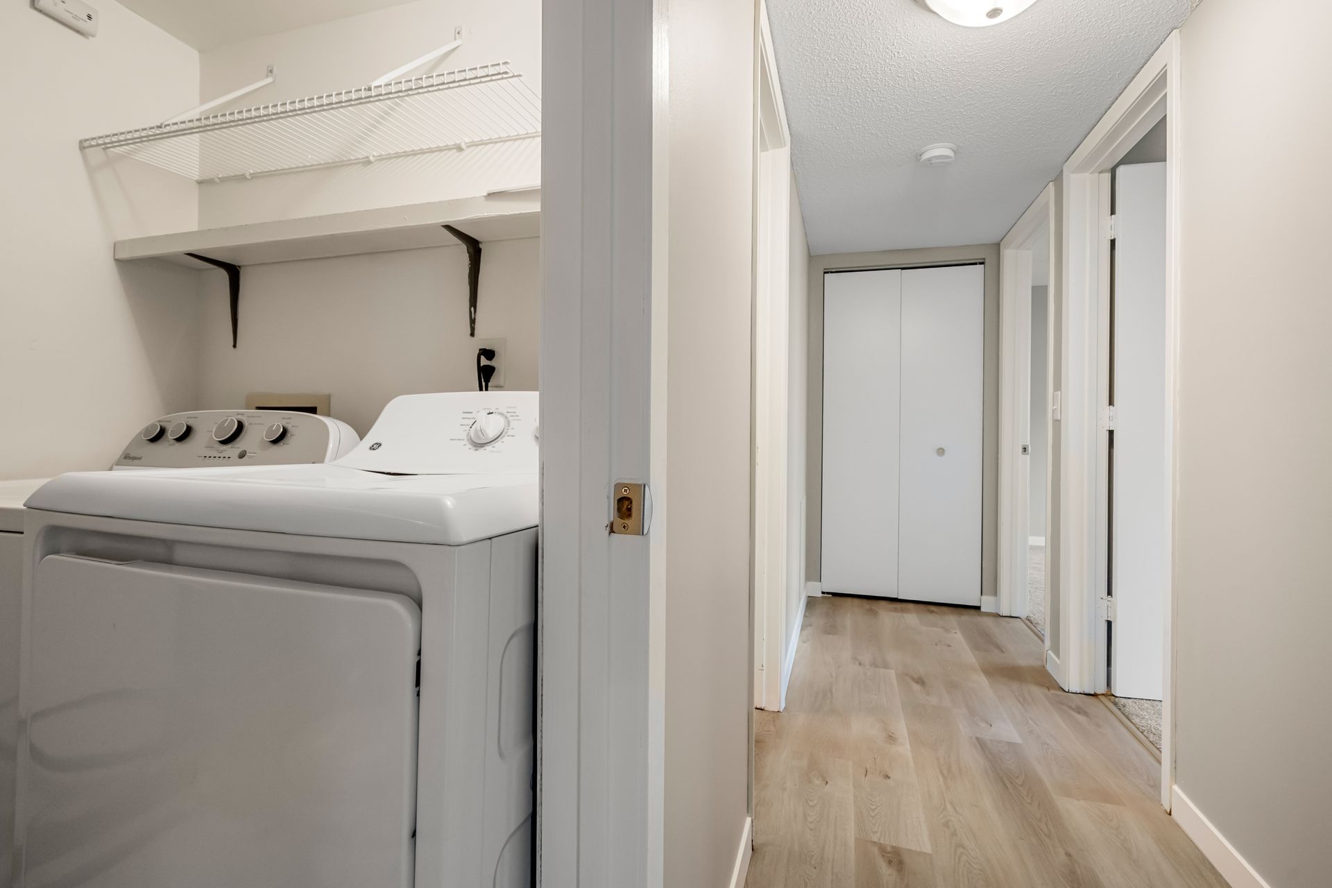 Laundry room and hallway with white washer and dryer, shelves above, and light wood flooring.