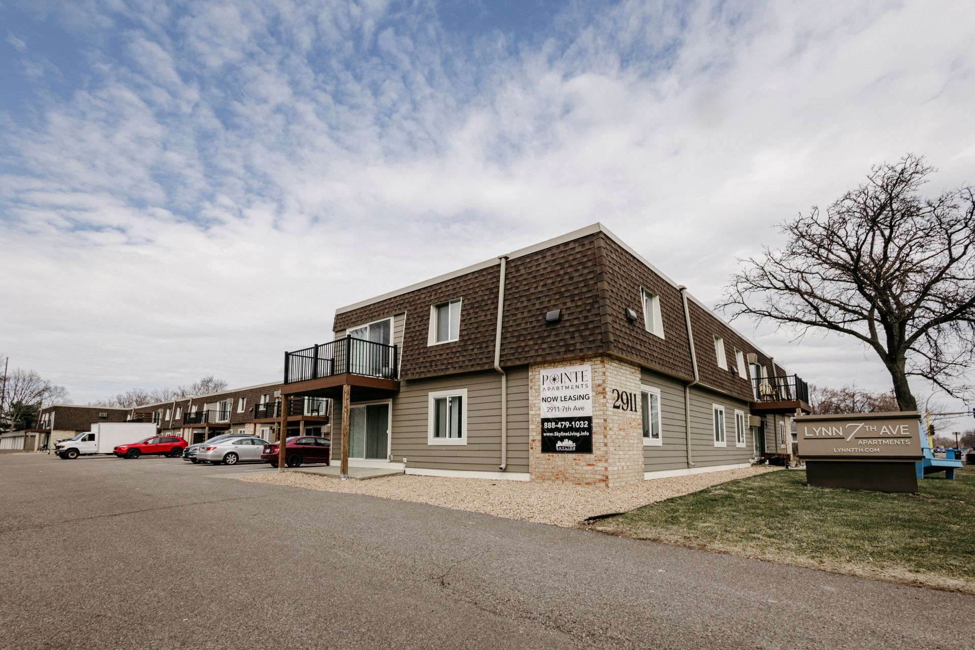 Apartment building with tan siding and balconies beside a gravel lot under a cloudy sky.