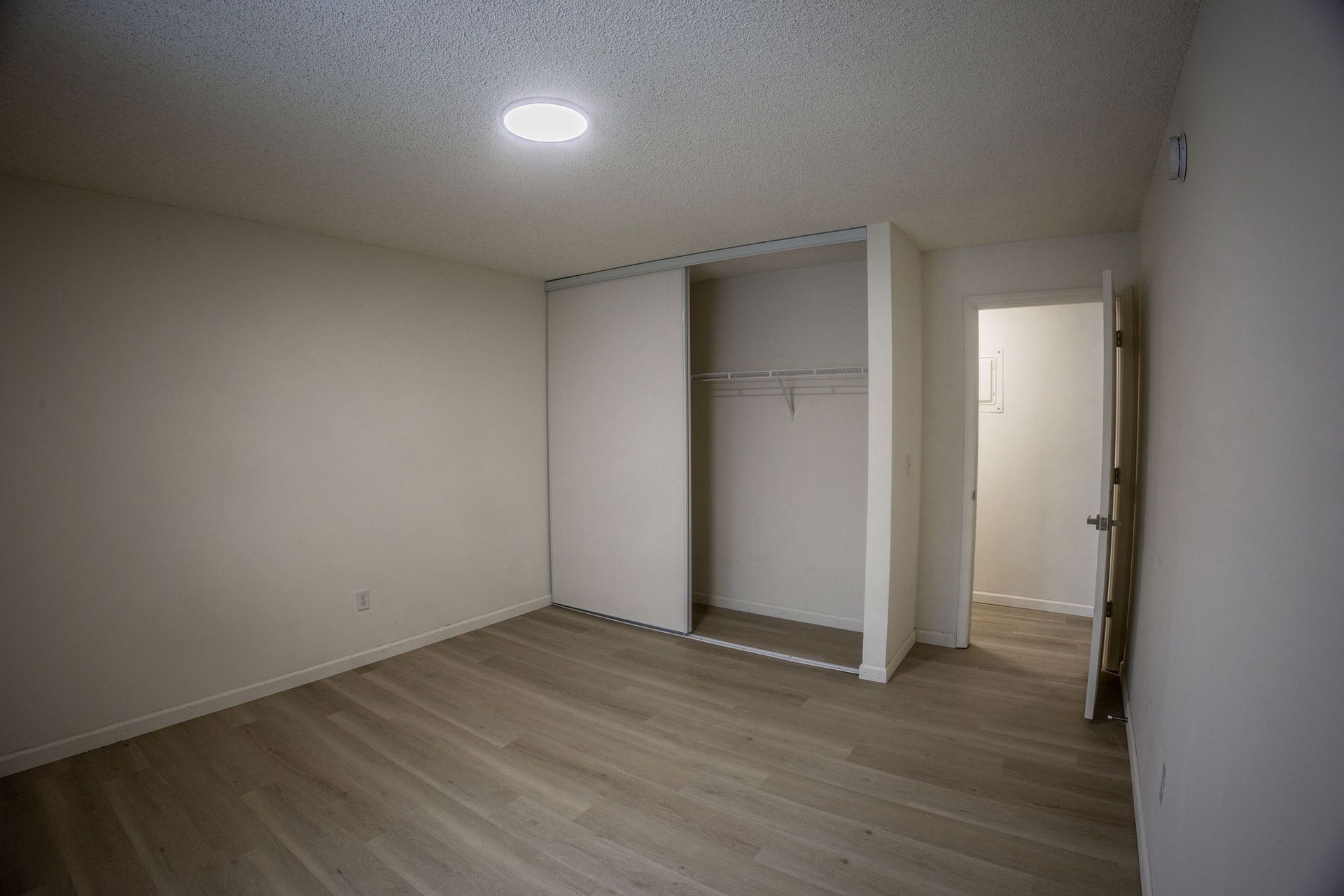 Empty bedroom with white walls, wood floor, open closet, and a doorway to a lit bathroom.