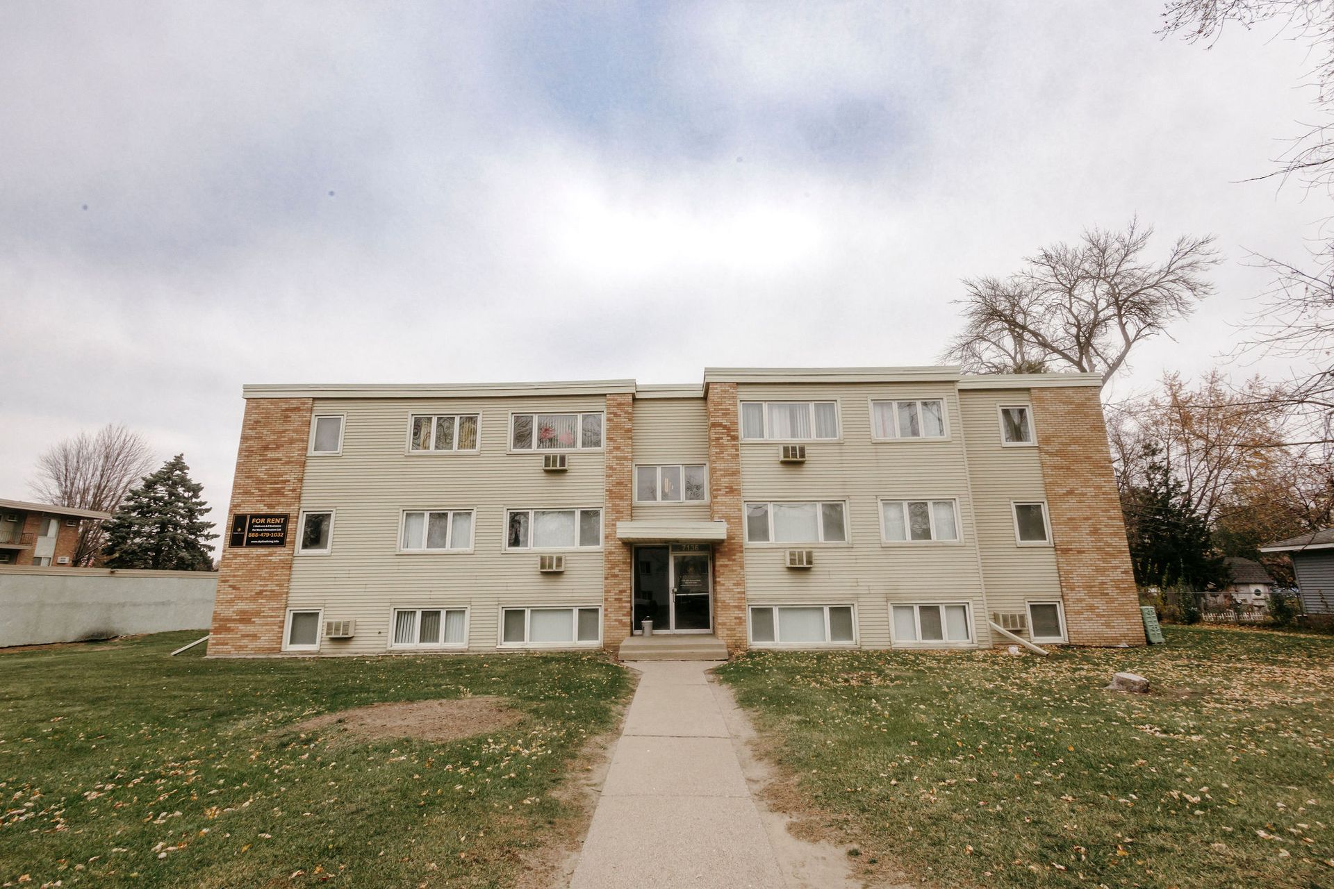 Three-story beige apartment building with a central entrance and a sidewalk leading to it.
