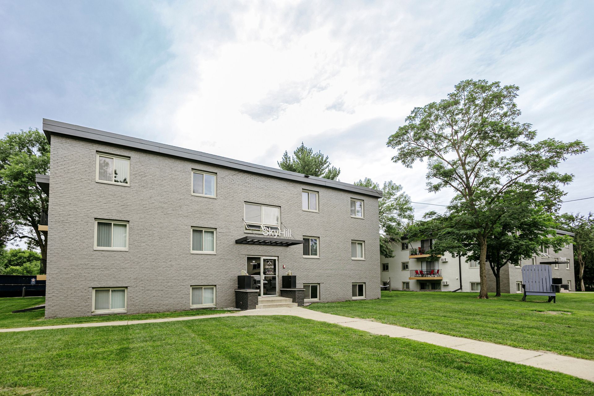 Brick apartment building with grassy lawn, sidewalk, and trees under a cloudy sky
