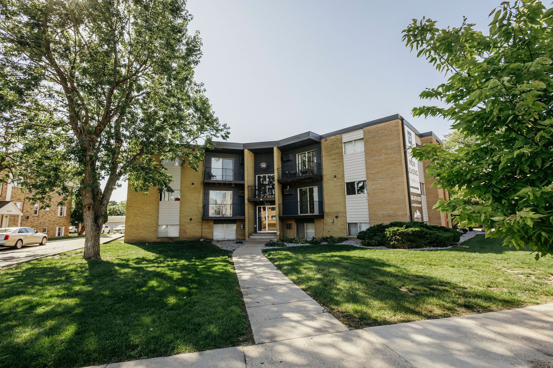 Apartment building with tan brick, black balconies, and a sidewalk through a grassy front lawn