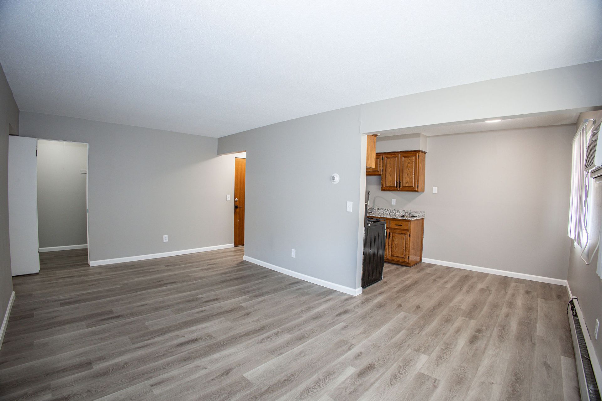Empty living room with gray wood floors, light walls, and a small open kitchen nook.
