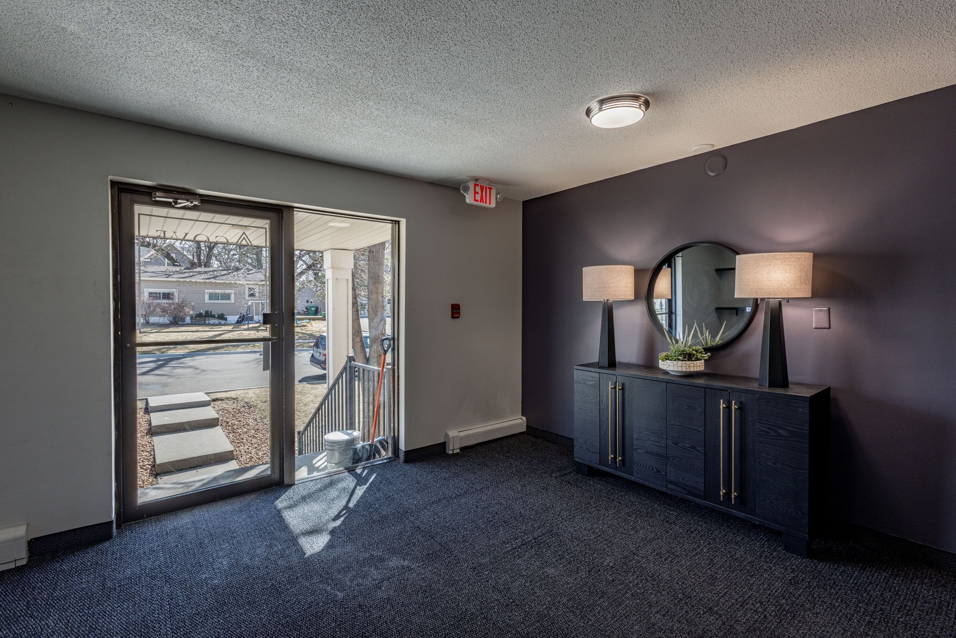 Empty room with dark carpet, purple wall, black console, lamps, and glass door to snowy outdoors