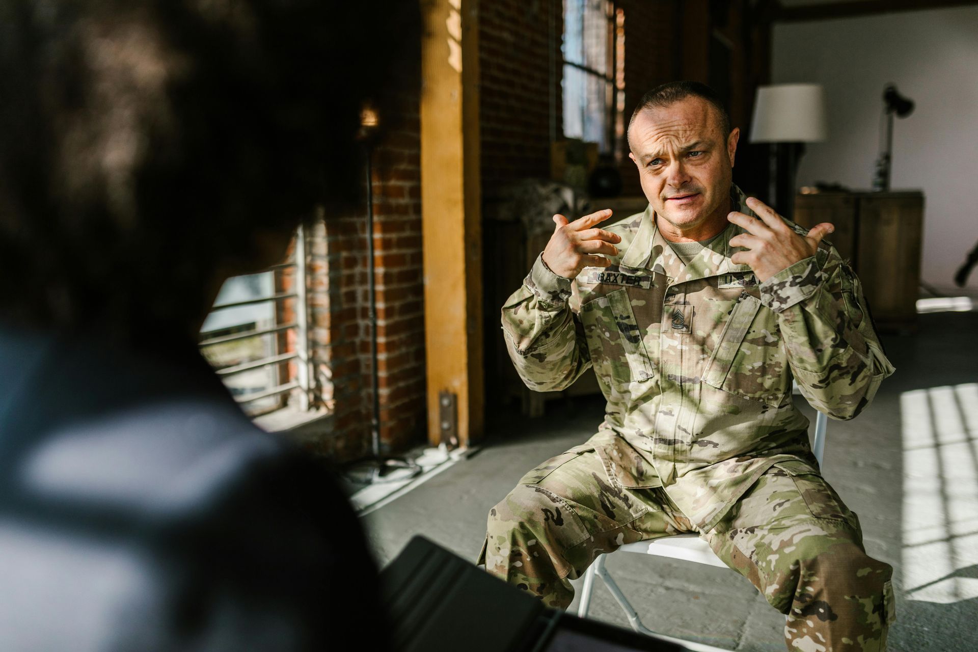 Man in military uniform speaking indoors, seated beside a window with brick walls and a lamp.