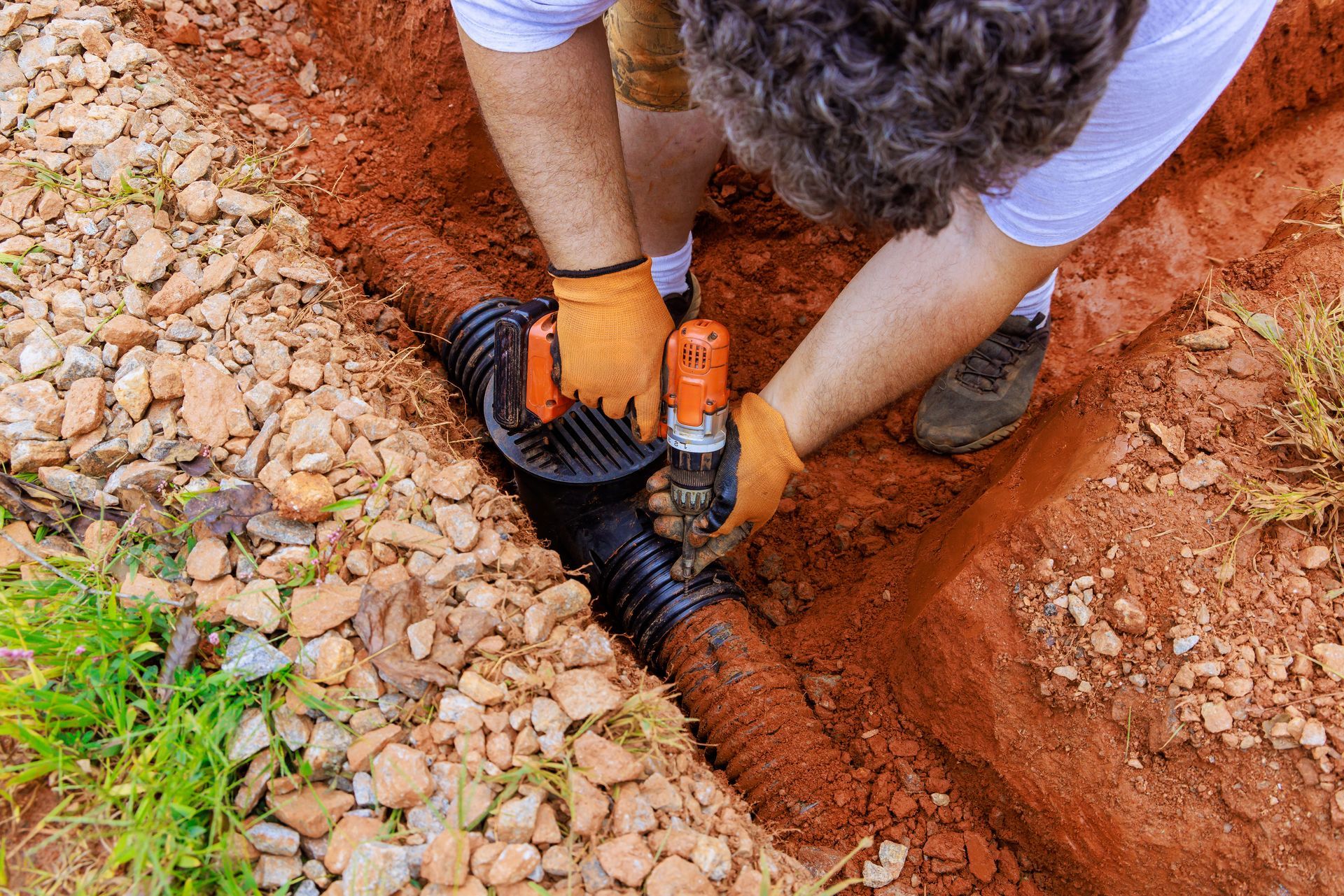 Person in gloves connects drainage pipe in a red dirt trench, beside gravel and plants.