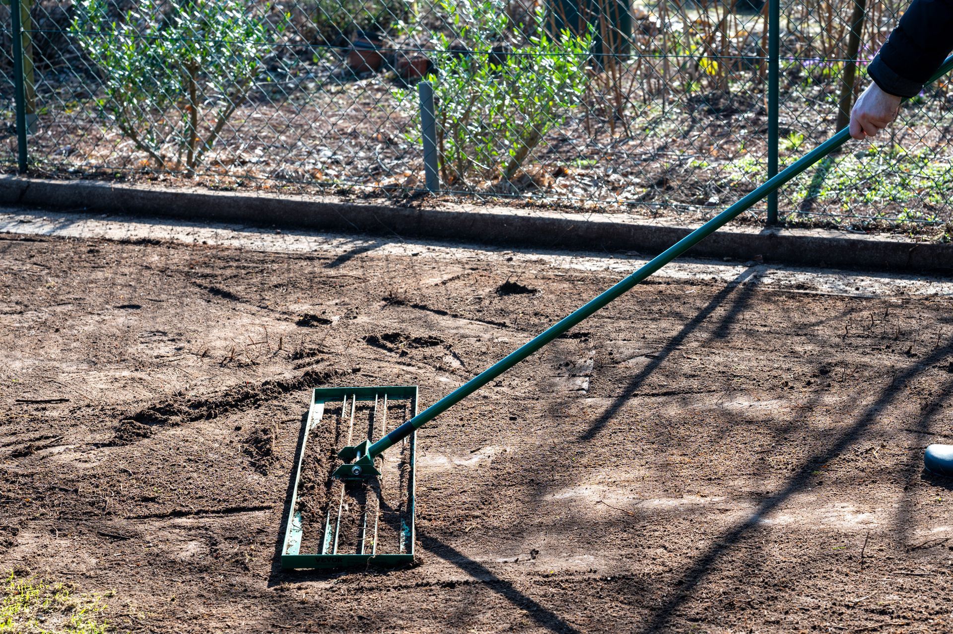 Person using a lawn leveler to smooth a garden bed. Green tool, brown soil, fence in background.