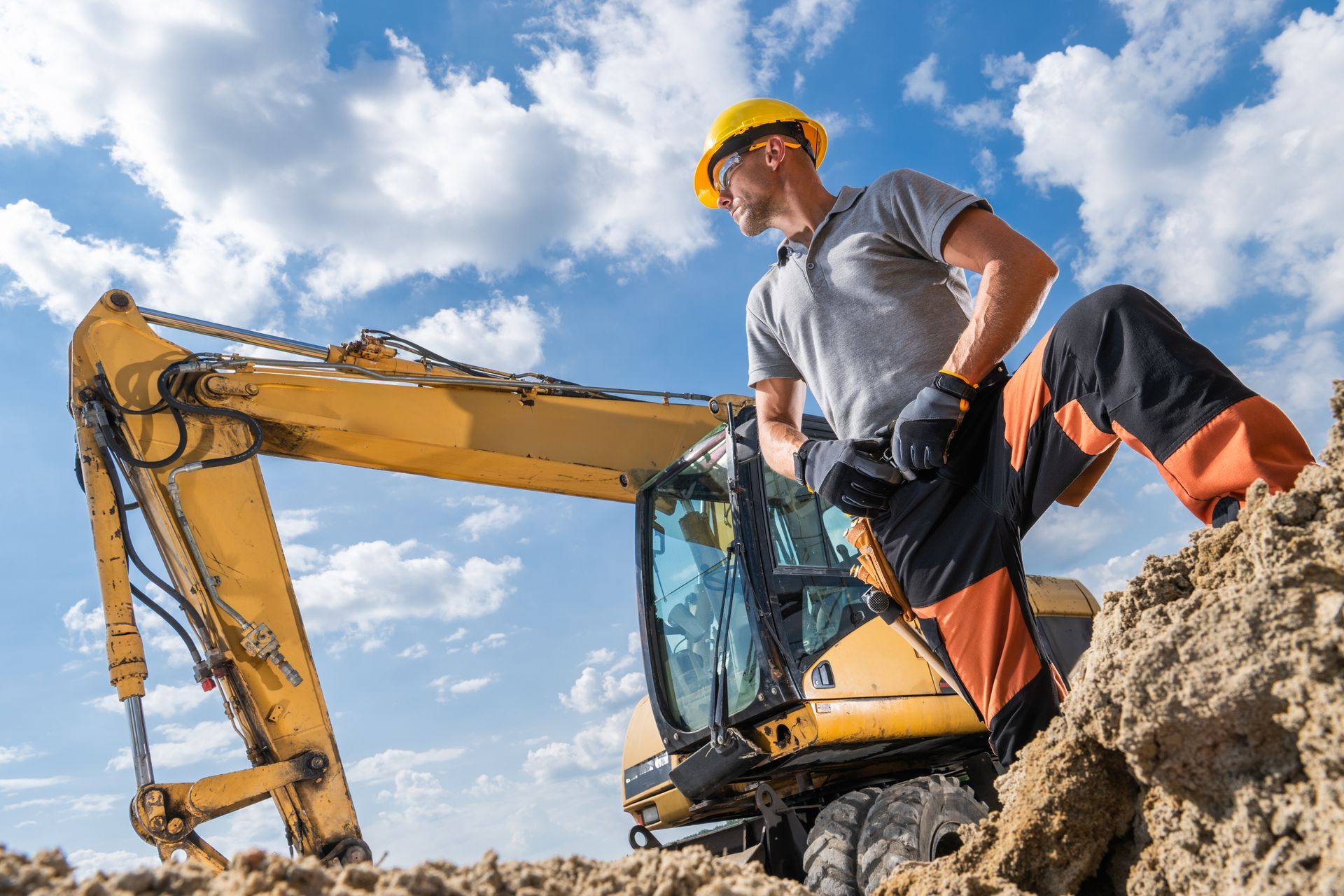 Construction worker in hard hat, leans on yellow excavator, sunny sky.