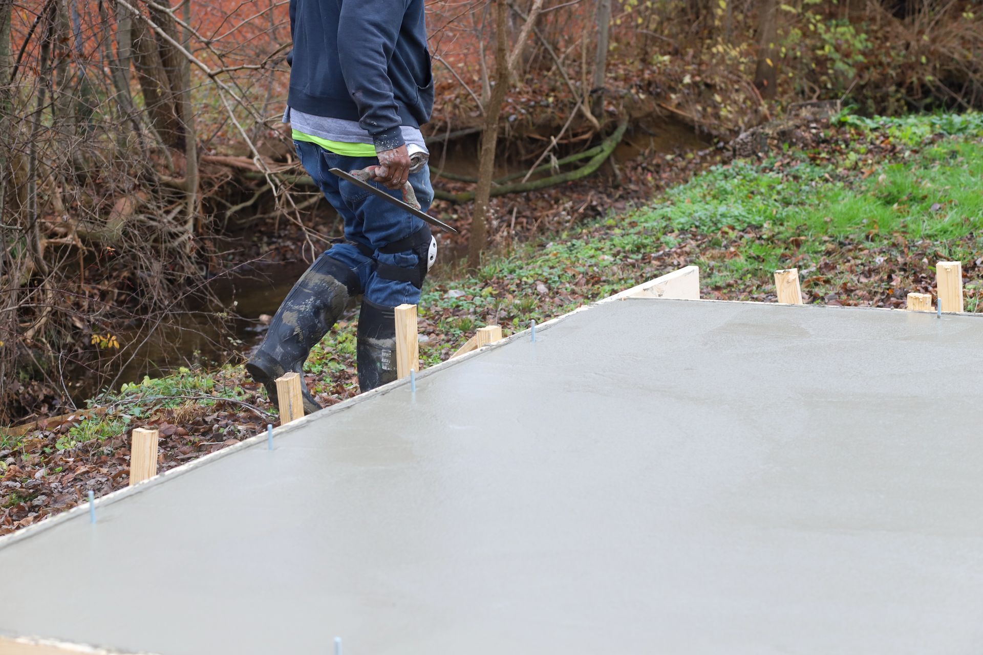 Man leveling wet concrete surface with a tool. Outdoors, wooden stakes in the foreground.
