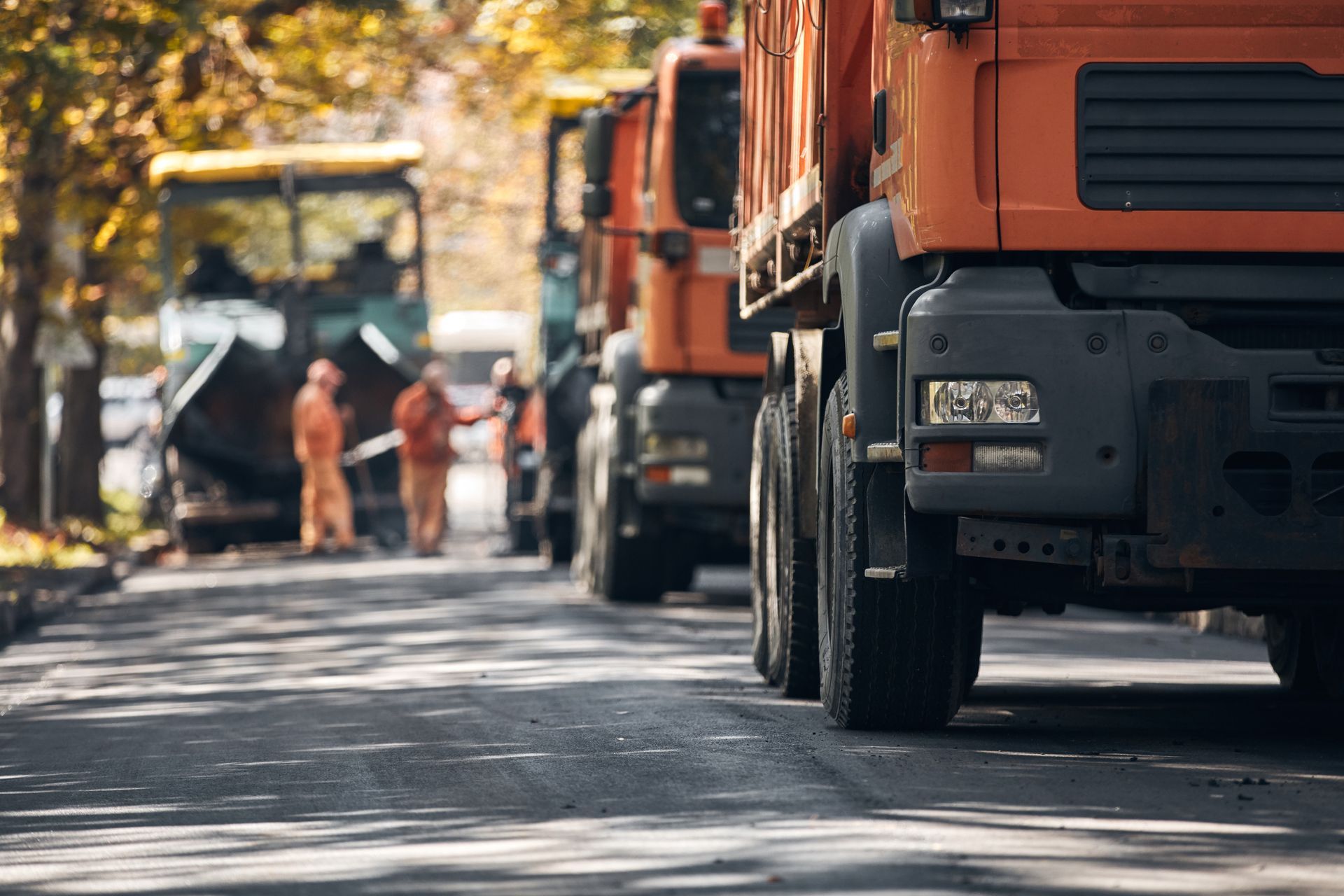 Road construction scene with orange dump trucks and workers on asphalt.