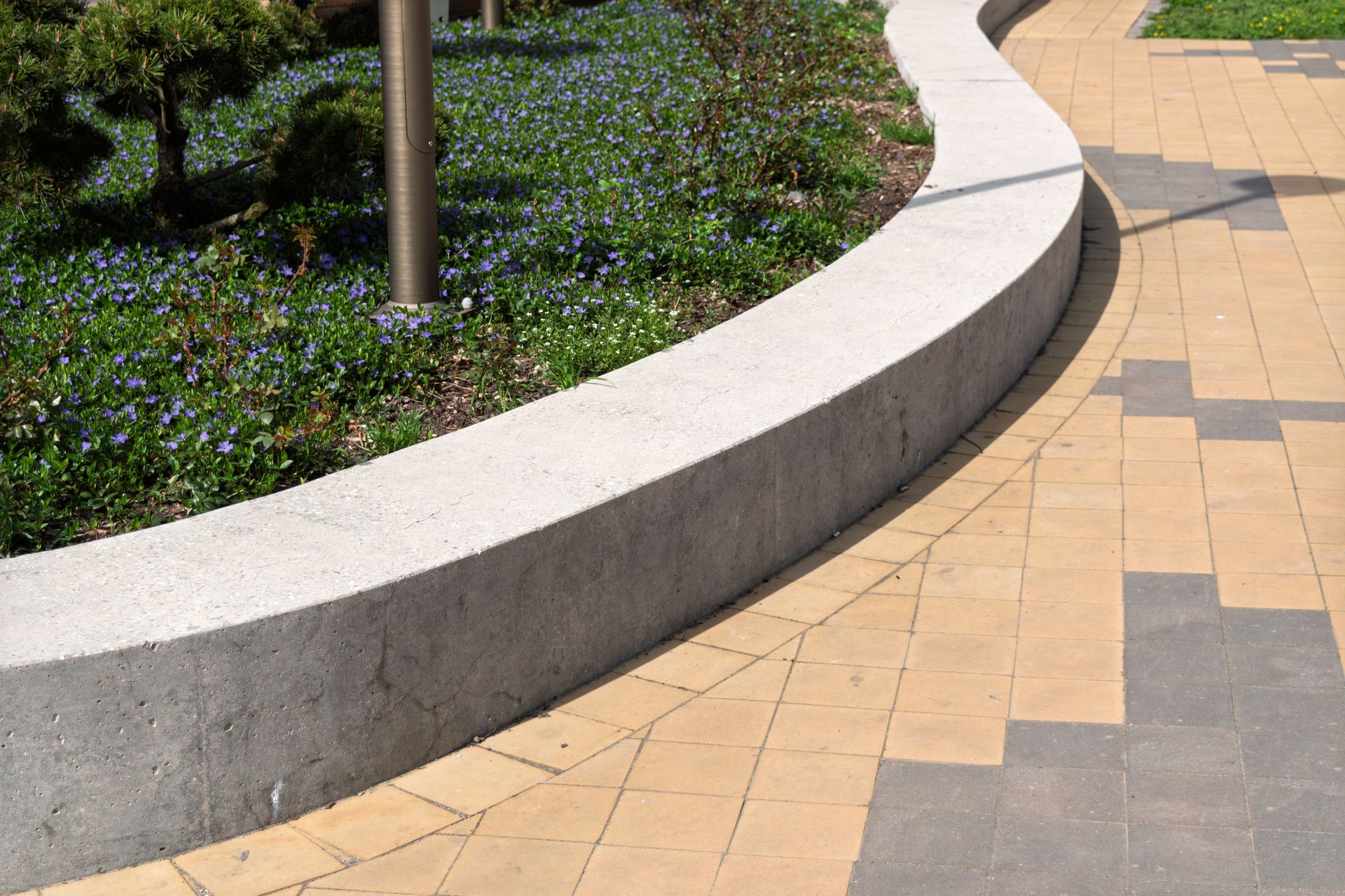 Curved concrete border separating a garden of blue flowers and paving stones in a park setting.