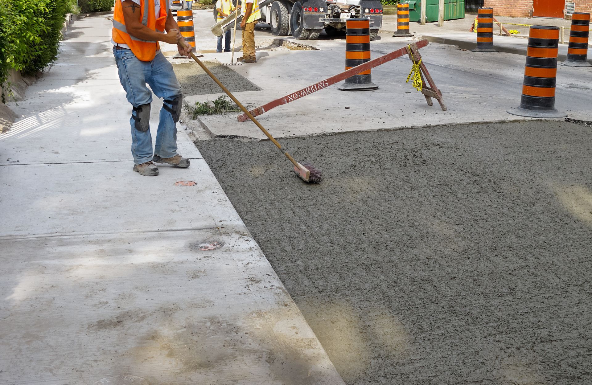 Construction worker leveling concrete with a screed on a road repair site, orange safety cones present.