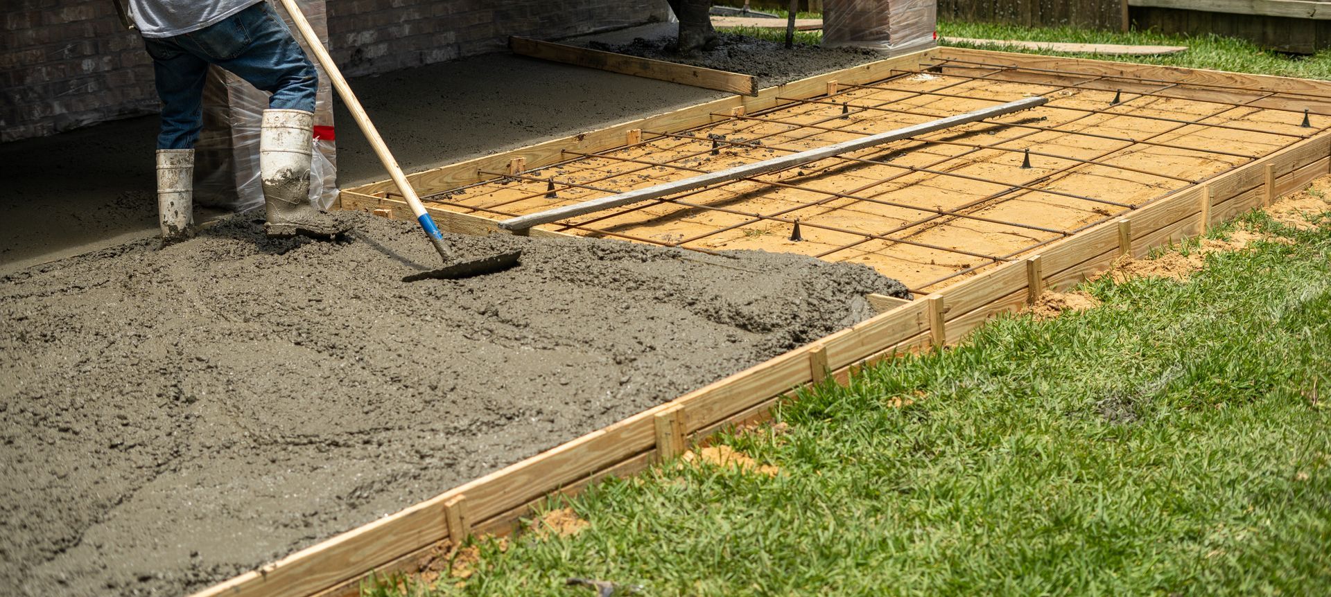 A worker spreading wet concrete with a tool. The concrete is within a wooden frame on grass.