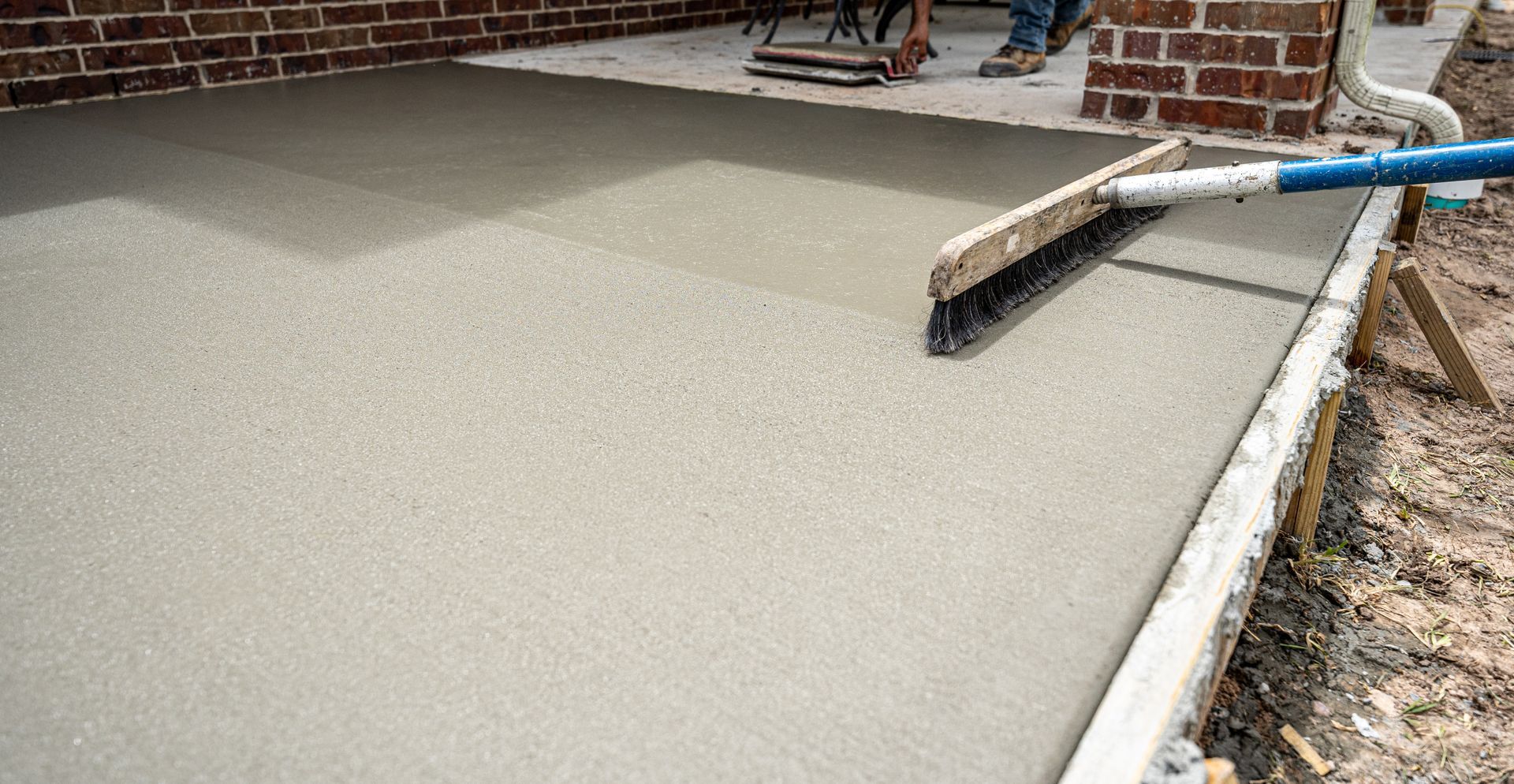 Freshly poured concrete being brushed smooth on a patio, near a brick wall and wooden forms.