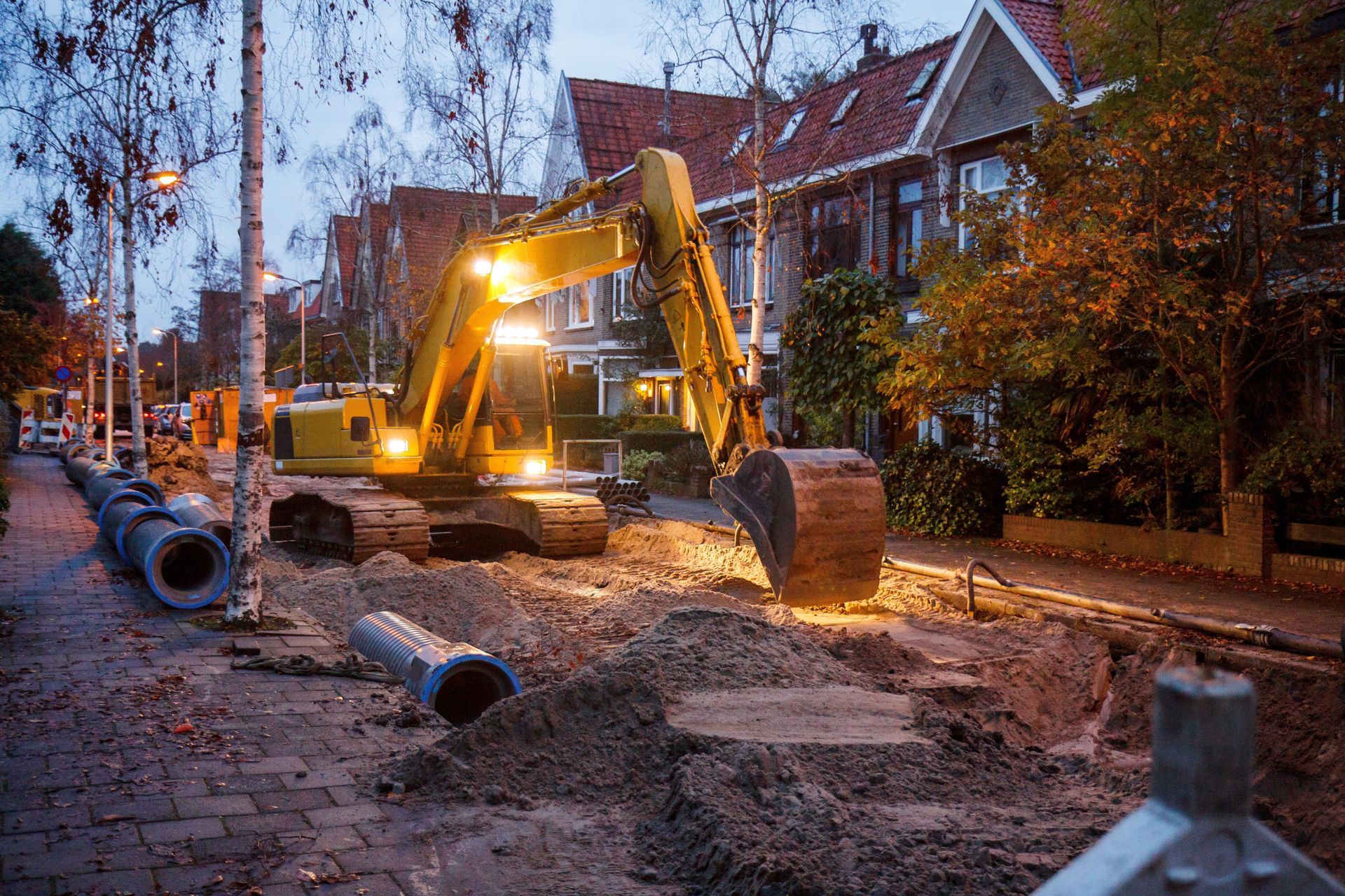 Excavator digging in residential street at dusk; pipes lie on sidewalk. Houses line the street.