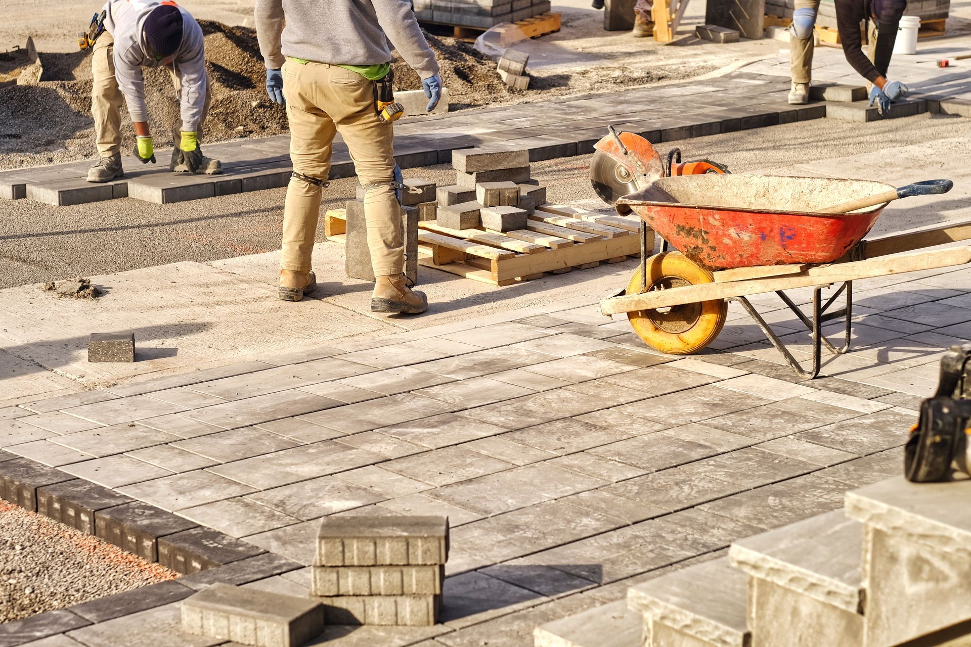 Construction workers laying pavers on a street. Wheelbarrow, tools, and stacked pavers are visible.