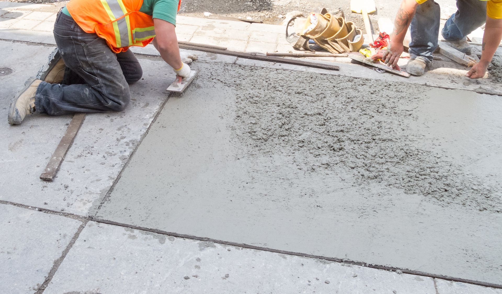 Workers smoothing wet concrete on a sidewalk, wearing orange vests.
