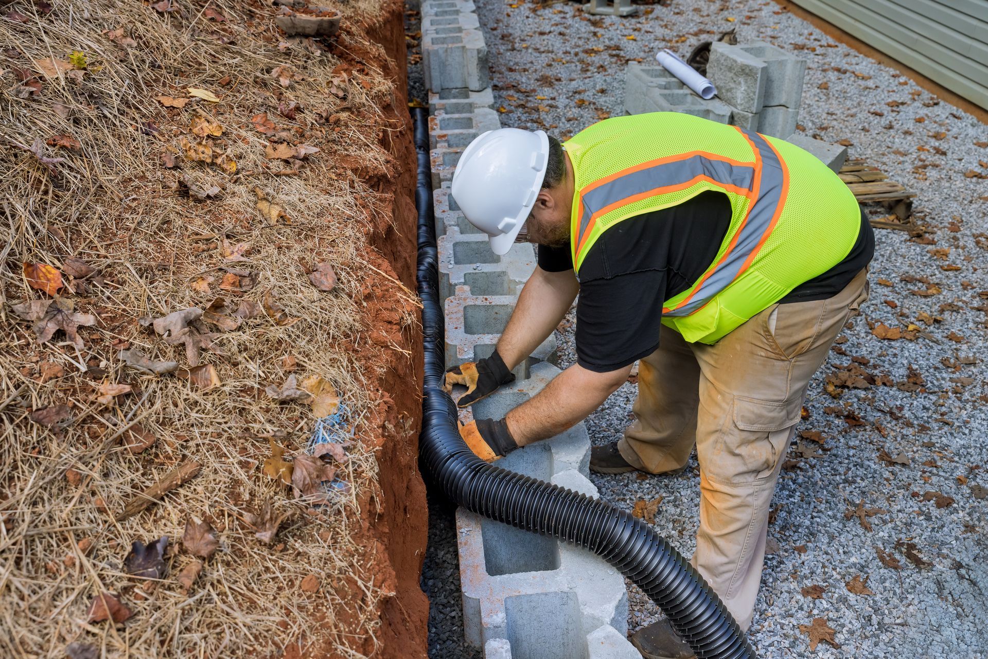 Construction worker installing black drainage pipe next to a cinder block wall in a trench.