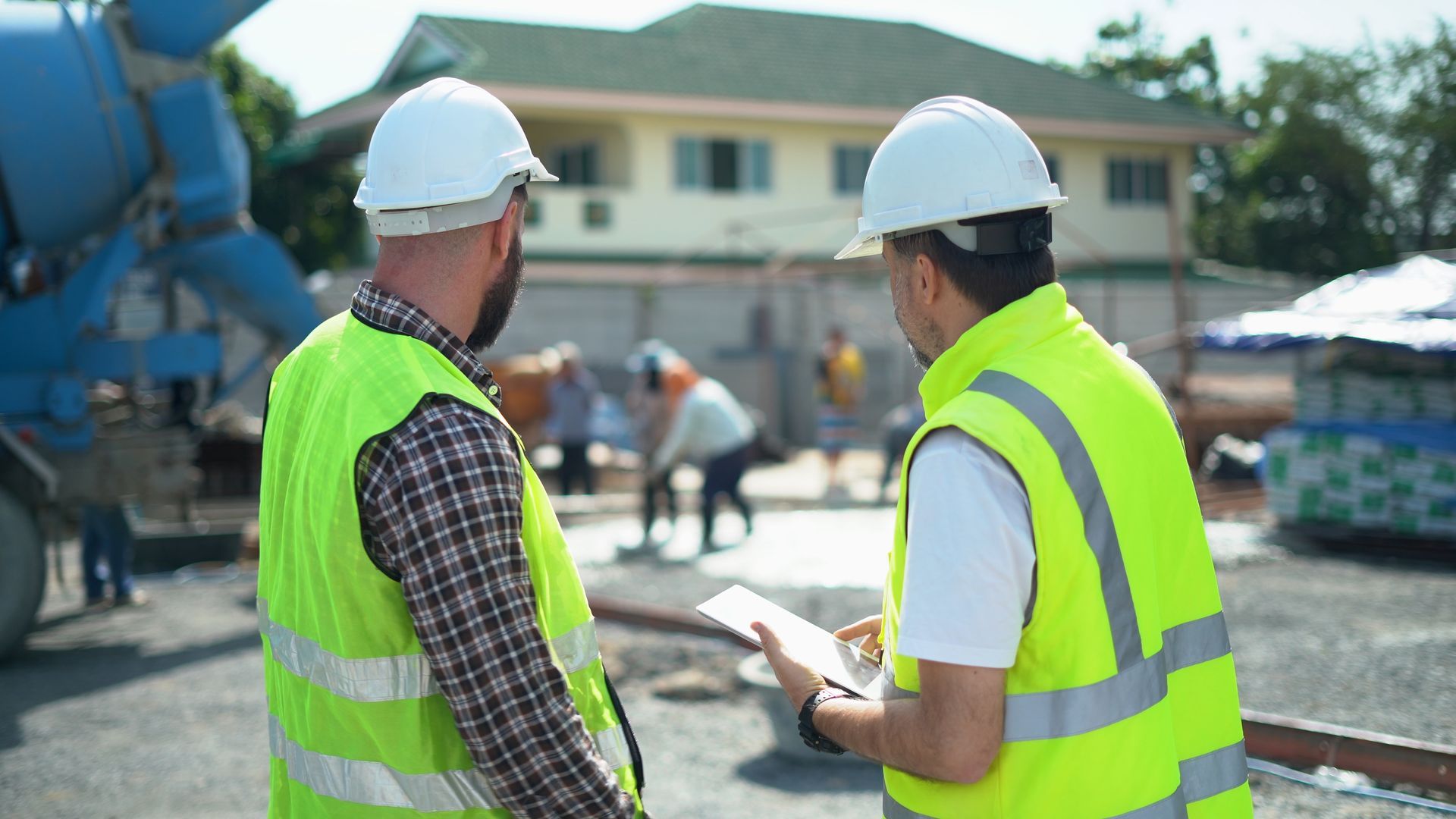 Two construction workers in vests and helmets review plans on site.
