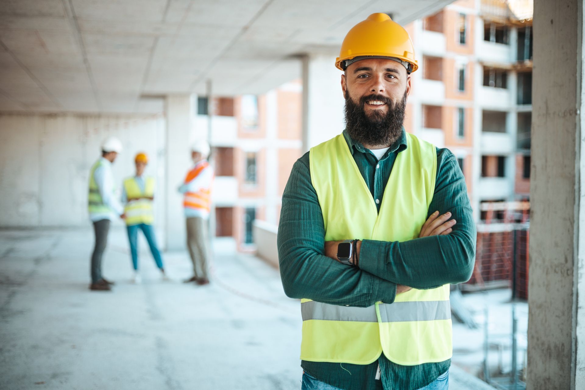Construction worker with arms crossed, smiling. Wearing yellow hard hat and vest. Other workers in the background.