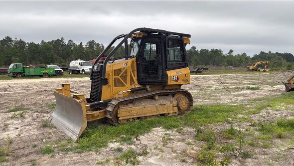 A yellow Caterpillar bulldozer sits on a dirt field with sparse grass, parked near other heavy machinery under a grey sky.
