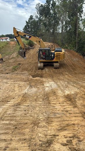 A yellow Caterpillar excavator sits on a sloped, cleared dirt construction site surrounded by trees.