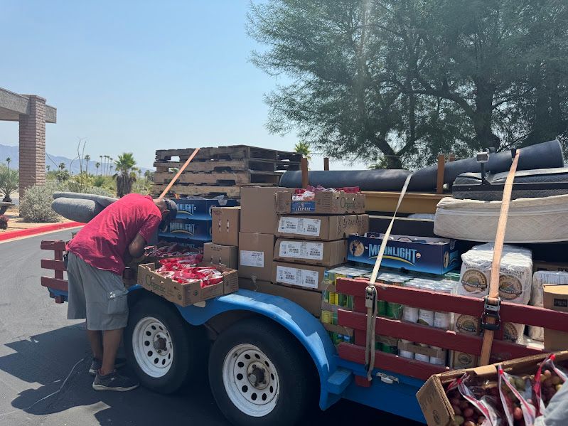 A person in a red shirt stands next to a blue trailer filled with stacked cardboard boxes, produce, and supplies.