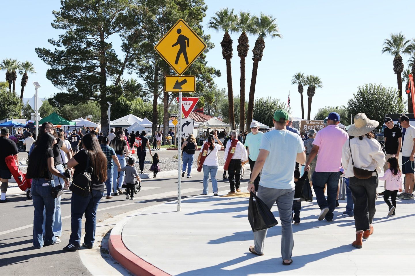 Pedestrians walk across a paved area near white vendor tents and palm trees, with a yellow pedestrian sign visible above.