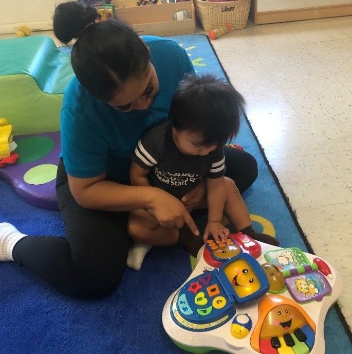 An adult sits on a blue rug and points to a colorful, interactive toy as a young child engages with it.