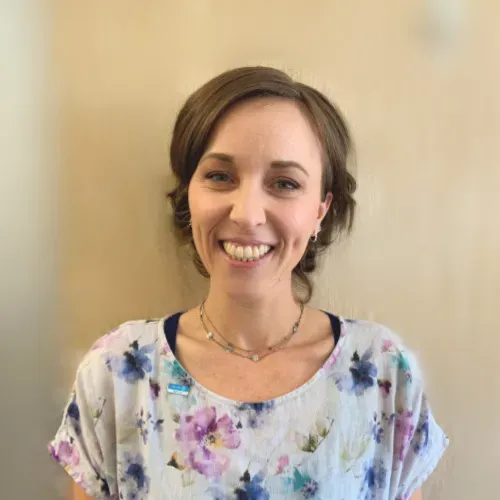 A smiling person with brown hair, wearing a floral print top and a double-layered necklace, stands against a beige wall.