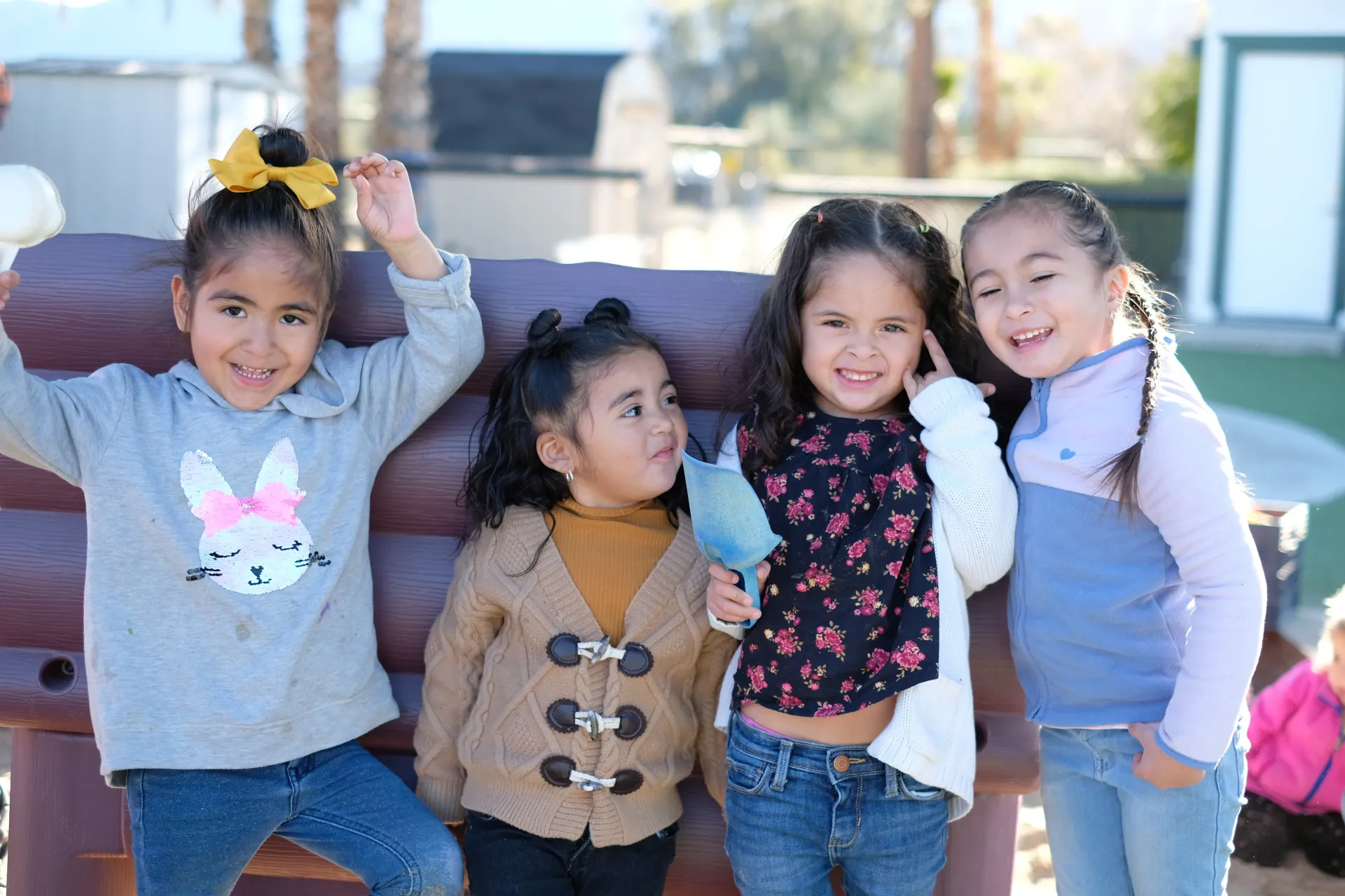 Four children standing together and smiling in front of a curved brown bench outdoors on a sunny day.