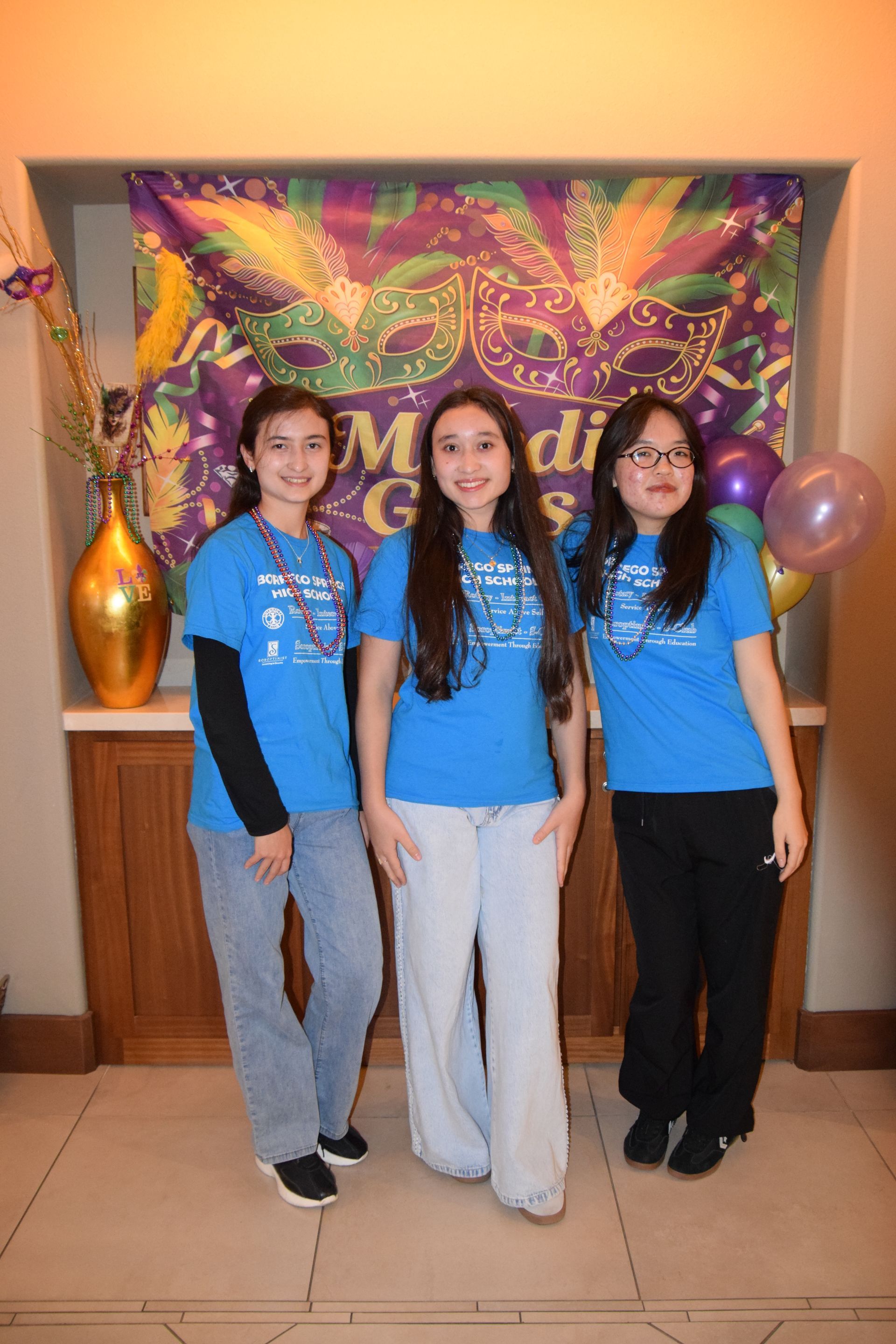 Three people in matching blue shirts pose together in front of a colorful Mardi Gras-themed backdrop and decorations.